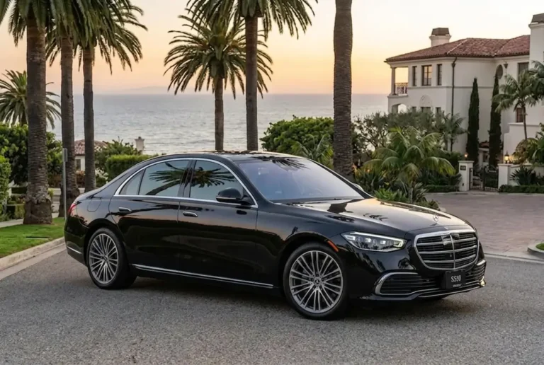 A black Mercedes-Benz S550 sedan parked on an asphalt road in front of a large, Mediterranean-style estate. The car is angled slightly toward the right, showcasing its front grille, headlights, and side profile. The background includes tall palm trees and a view of the ocean under a clear sky at sunset.